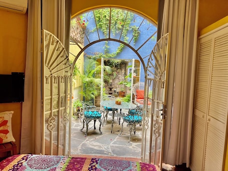 A room with a view of a courtyard, a bed with a colorful blanket, and a TV on the wall.