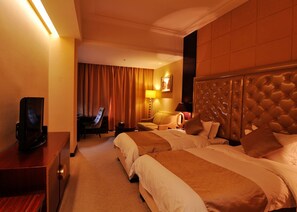 Ein Hotelzimmer mit zwei Betten, einem Schreibtisch mit Fernseher, einem Sessel, einer Lampe und einem großen Kopfteil.