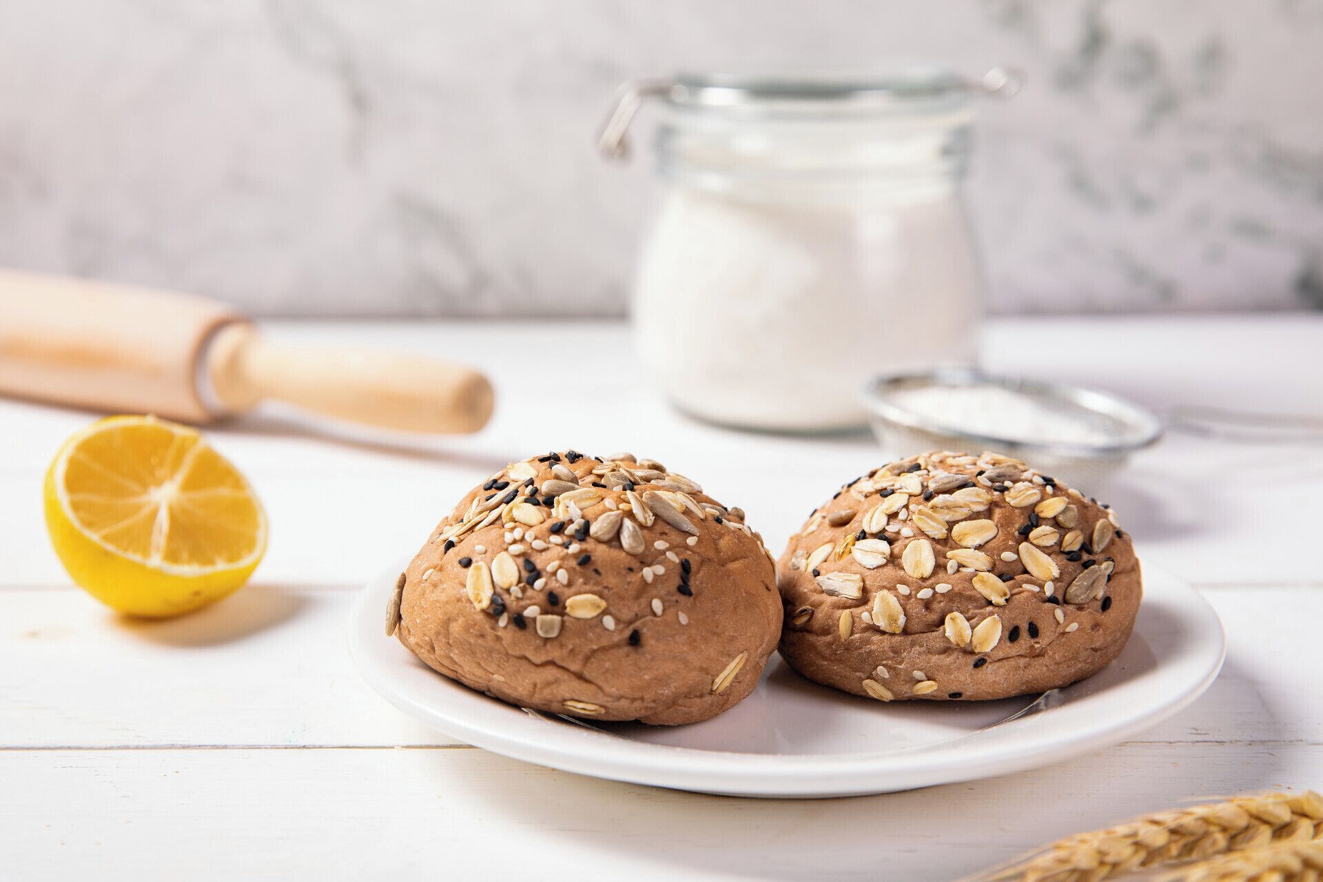 Two round, seeded breads on a white plate with a glass jar of milk and a rolling pin in the background.