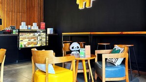 Interior de una cafetería con muebles de madera, una vitrina con productos alimenticios y un gran símbolo de numeral en la pared.