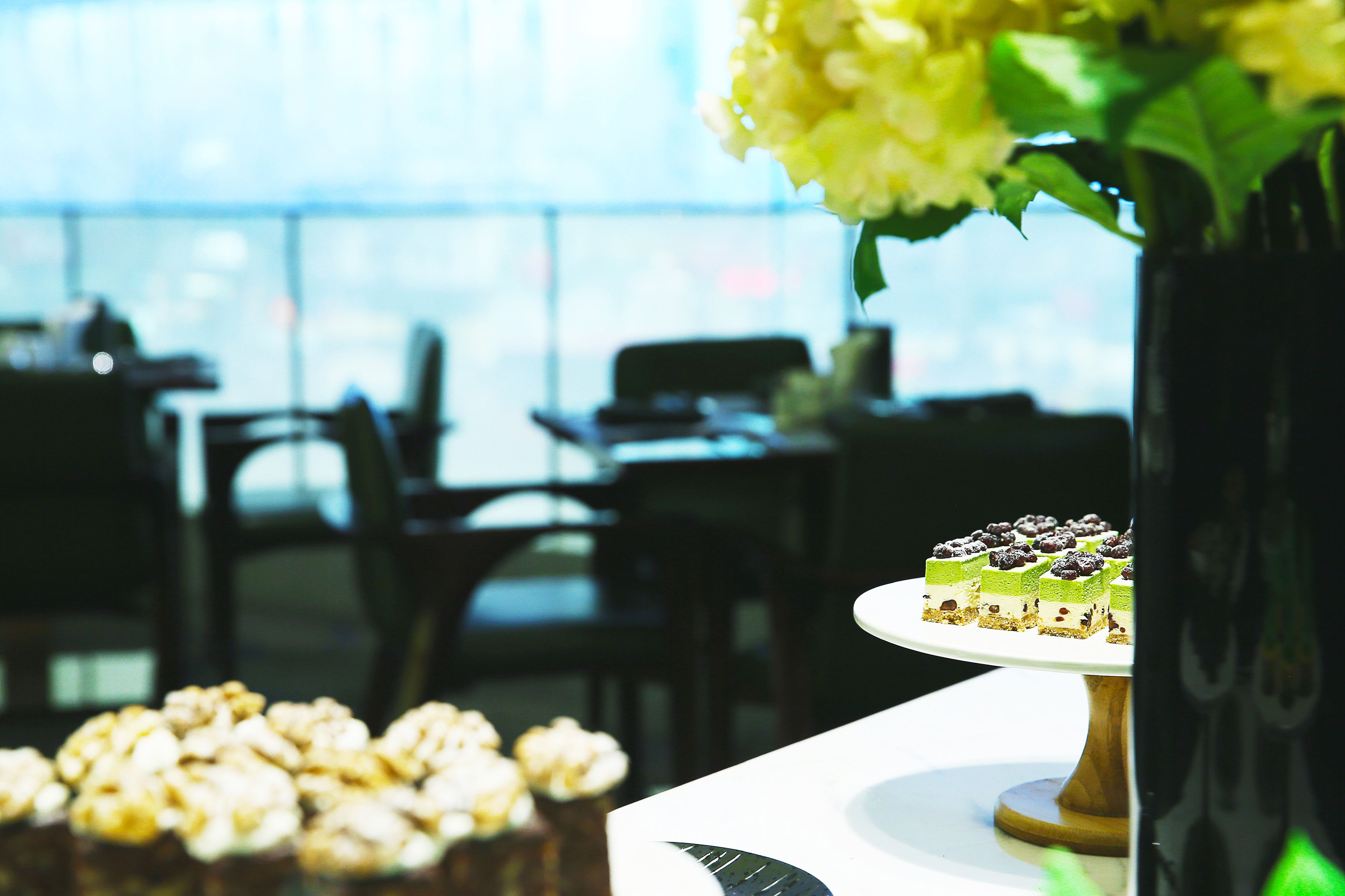 Une table blanche est dressée dans une pièce lumineuse, avec une grande fenêtre et un vase rempli de fleurs jaunes. On y trouve une sélection de desserts, notamment un gâteau et des cupcakes.
