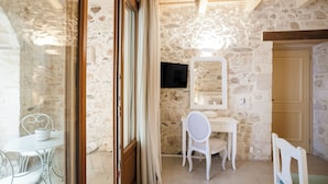 A room with stone walls, a wooden ceiling, a white desk, a chair, a television mounted on the wall, and a door. - Z boutique Hotel (Mylopotamos)