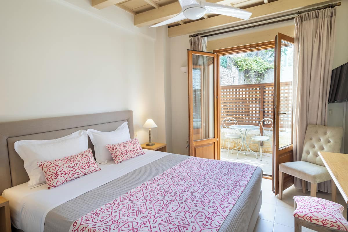 Habitación de hotel con cama, mesita de noche, lámpara, ventilador de techo y puerta corrediza de cristal a un espacio exterior con mesa y sillas.