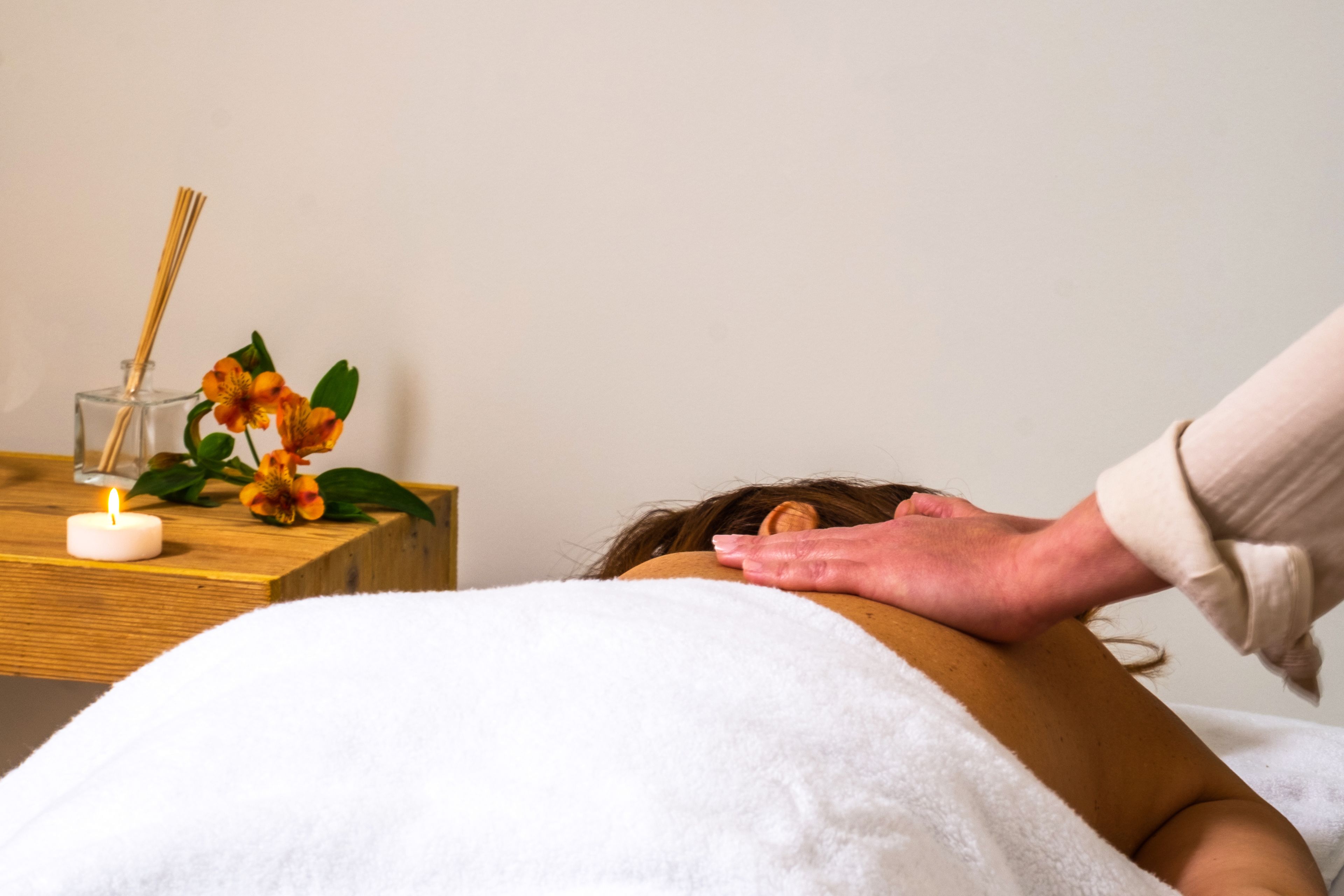 Eine Massageanwendung in einem Raum mit einem Holztischchen, einer Kerze und Blumen.