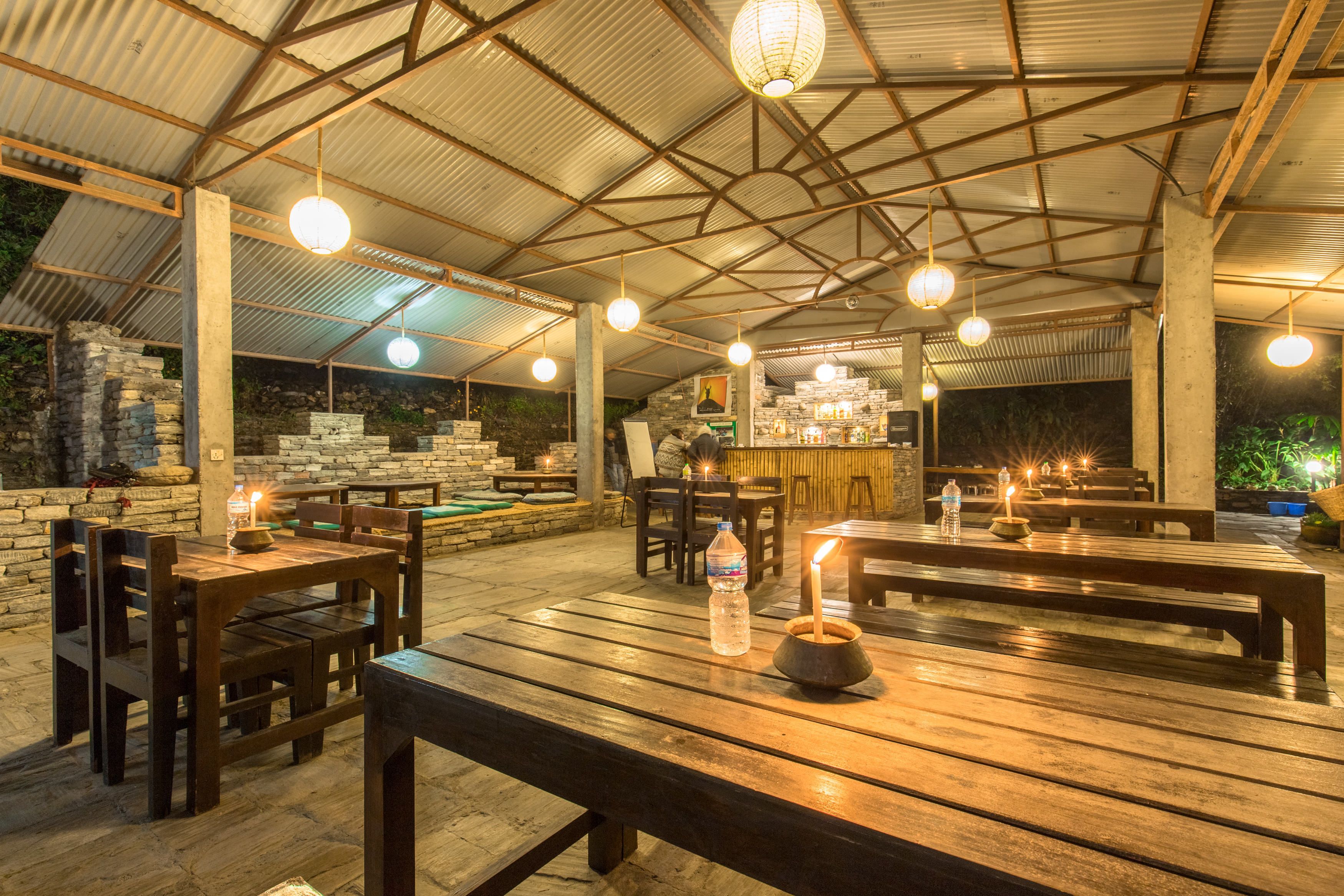 Un área de comedor al aire libre con mesas y bancos de madera, velas y luces colgantes.