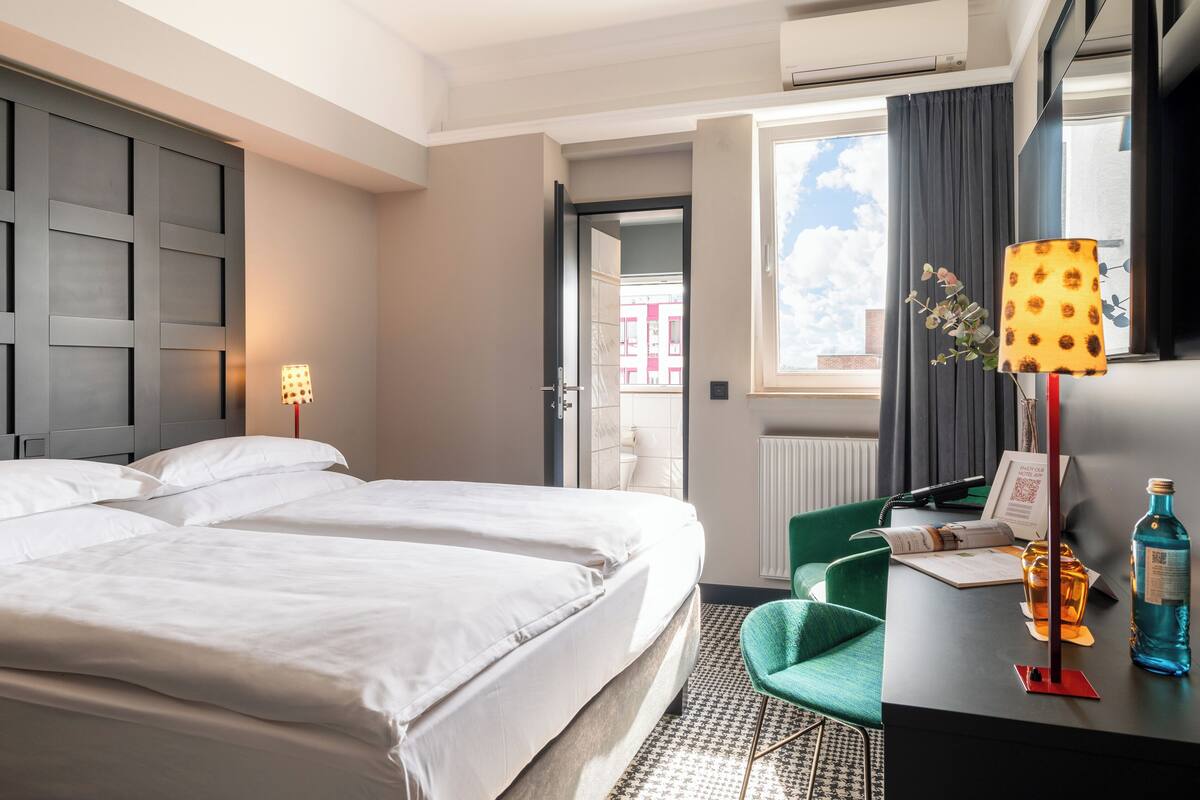 A hotel room with two beds, a desk with a lamp, a bottle, and a chair.