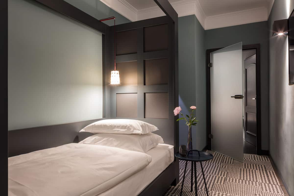 A modern hotel room with a neatly made bed, a bedside table with a vase of flowers, and a wall-mounted light fixture.