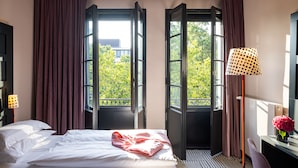 A hotel room with a bed, a desk, a lamp, and a view of trees through open windows. - The Midtown Hotel by The New Yorker (Cologne)