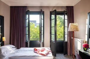 Quarto de hotel com cama, escrivaninha, luminária e vista para árvores através de janelas abertas.