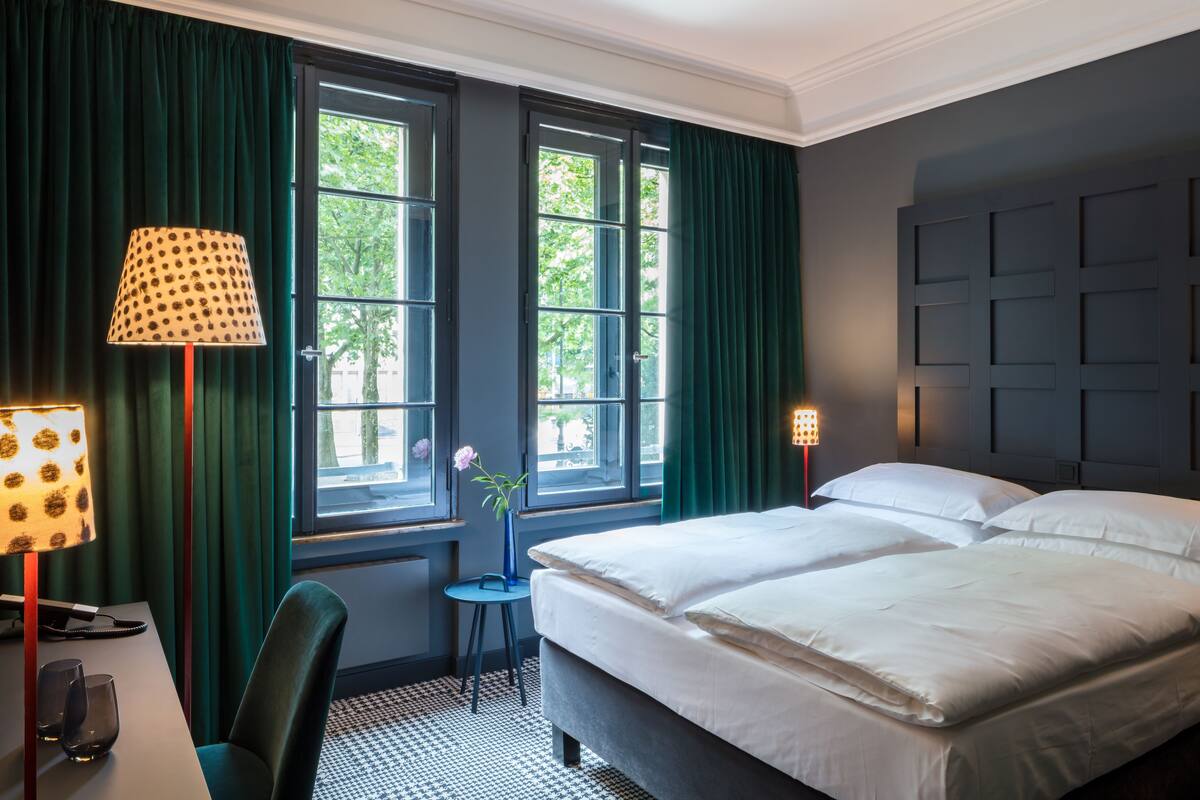 A hotel room with a large bed, two bedside lamps, a desk with a lamp, and a view of the outdoors through two windows.