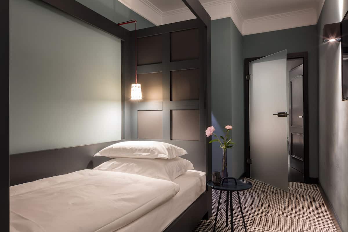 A hotel room with a bed, bedside table, vase with flowers, and a door leading to another room.