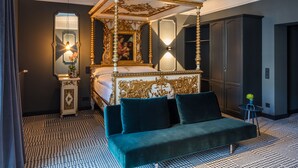 A four-poster bed with a gold and white ornate headboard, a green sofa, a small table with a plant, and a mirror on the wall. - The Midtown Hotel by The New Yorker (Cologne)