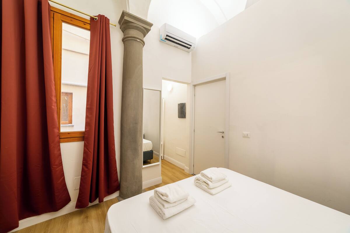 Une chambre d’hôtel avec un lit, une fenêtre avec des rideaux rouges, un appareil de climatisation et une porte donnant sur une salle de bain.
