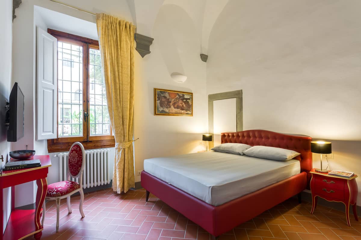 Une chambre avec un lit rouge, une table de chevet rouge, une chaise rouge, une fenêtre avec des rideaux, un radiateur et un tableau accroché au mur.