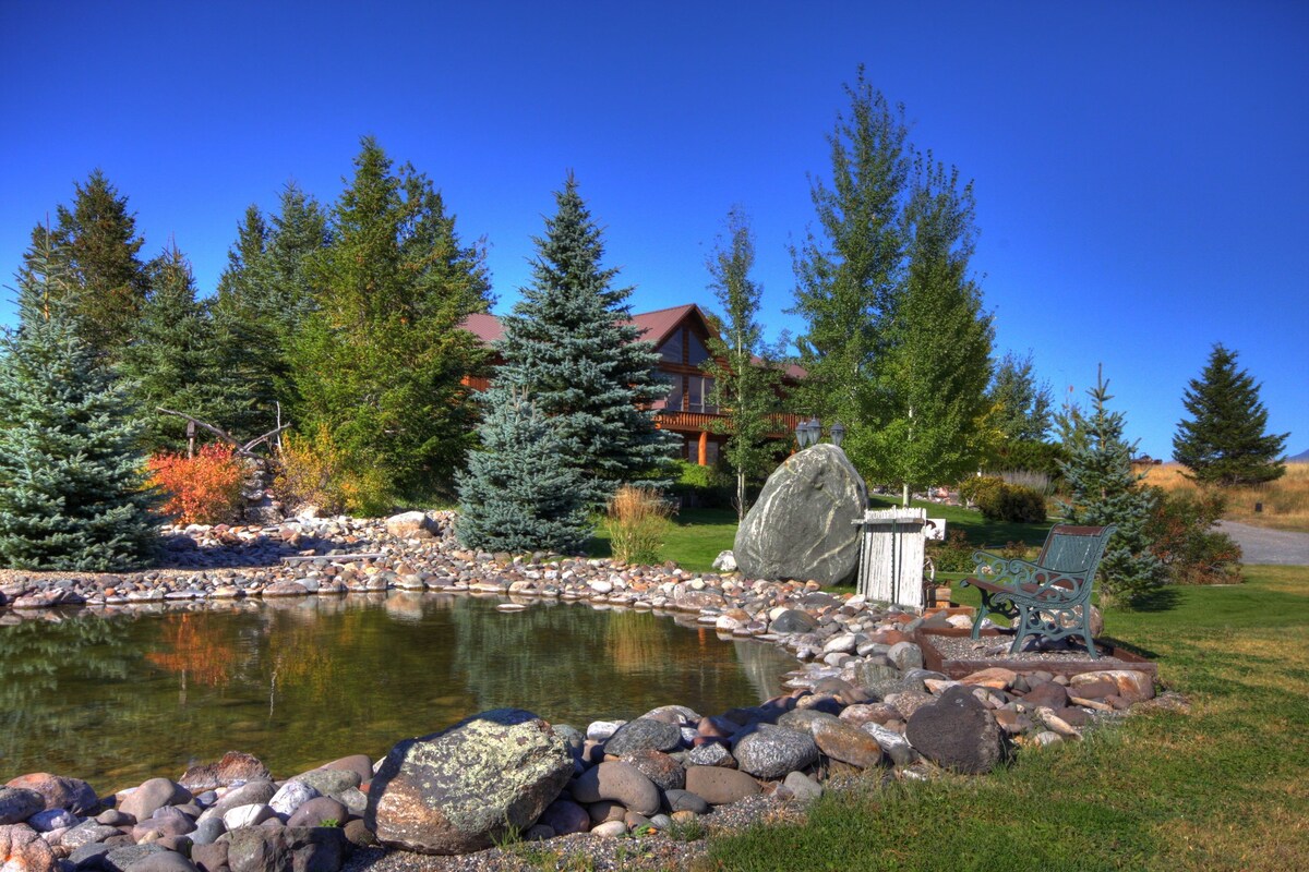 A pond surrounded by rocks and trees, with a wooden bench and a house in the background.