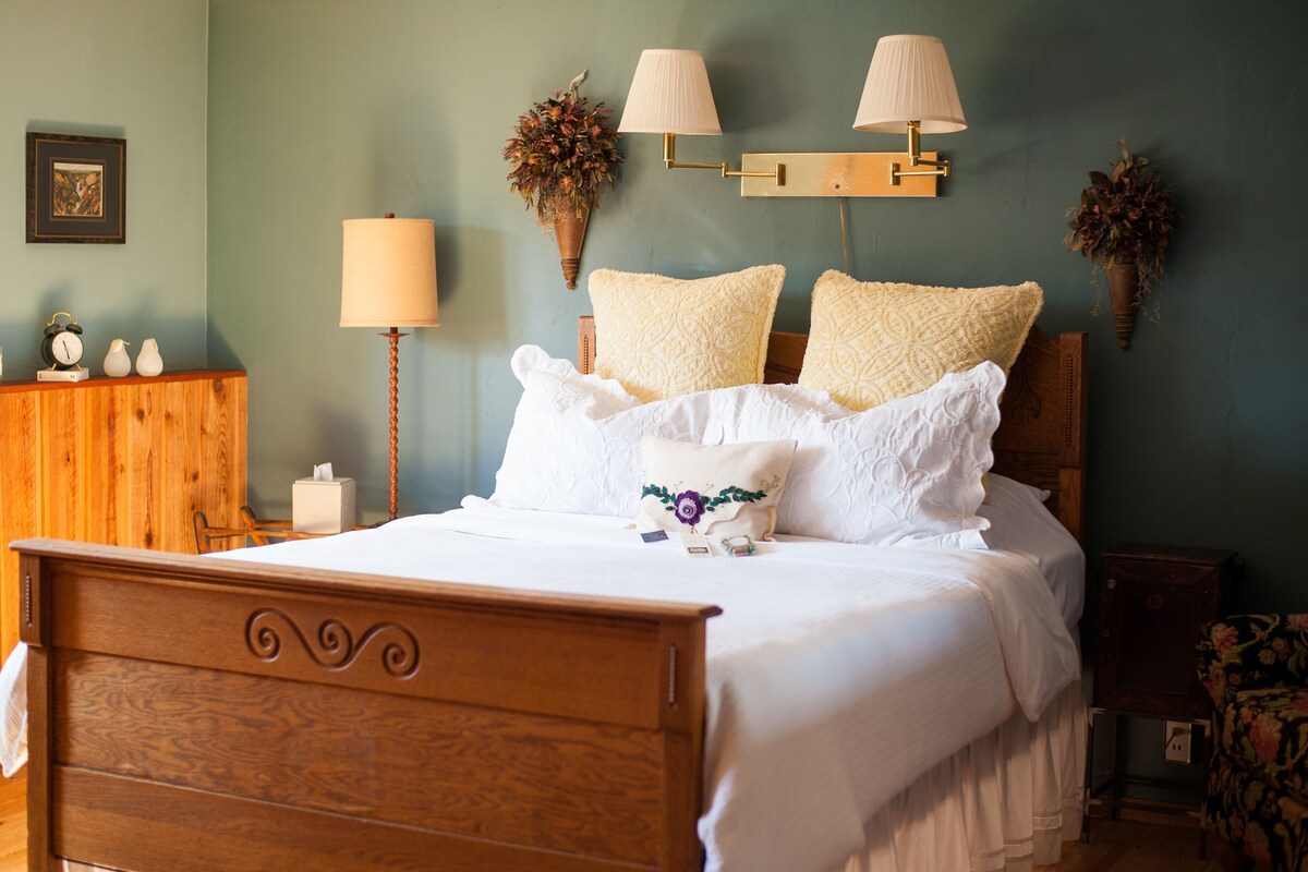 A neatly made bed with white linens and decorative pillows, a wooden headboard, and a bedside table with a lamp and a vase of flowers.