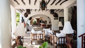 A spacious outdoor seating area with wooden furniture and hanging lanterns, leading to an archway and a view of the interior. - Scorpio Villas Resort (Malindi)