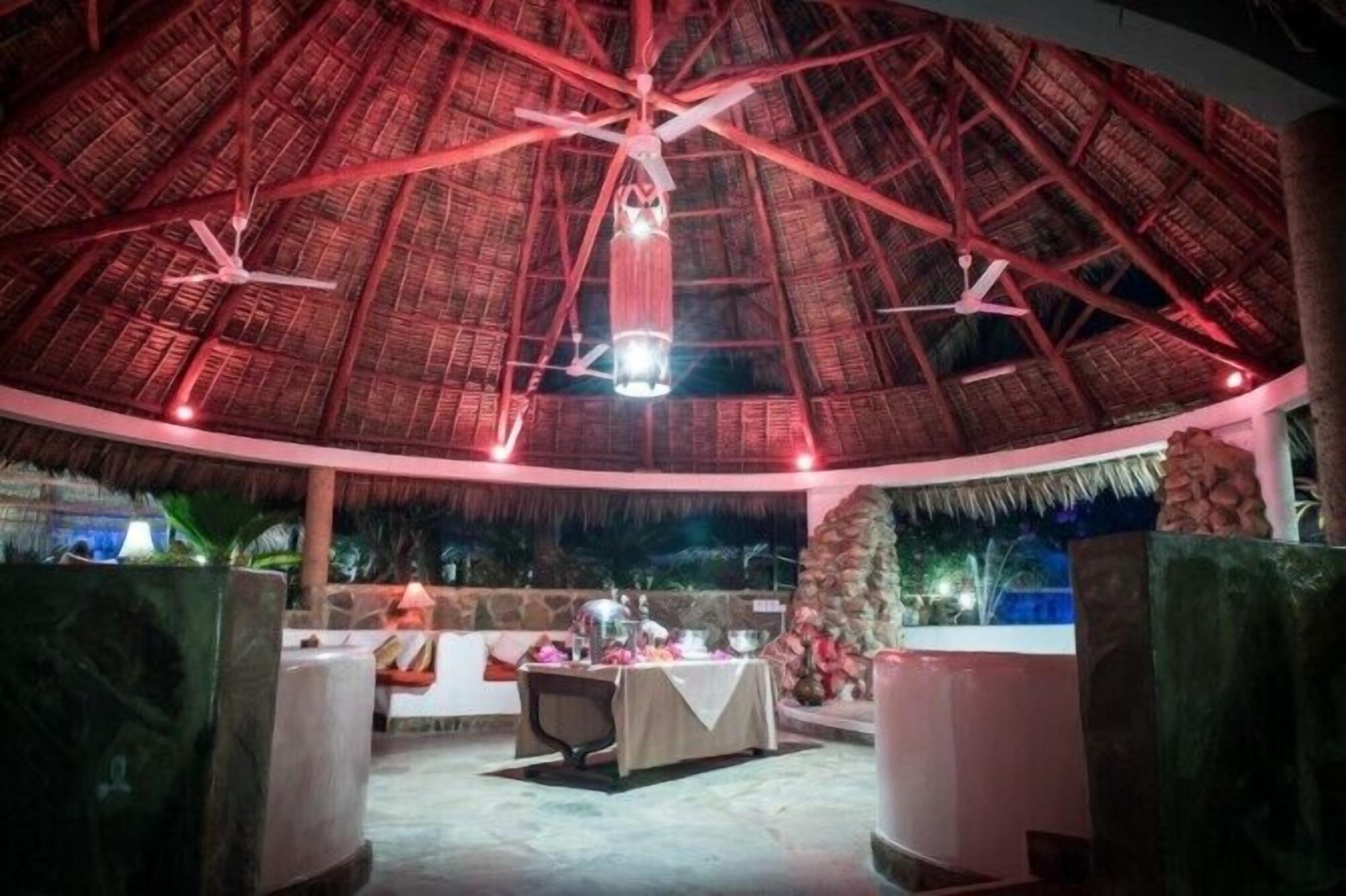 Un comedor interior con techo de paja, una mesa central dispuesta para comer, y un muro de piedra al fondo.