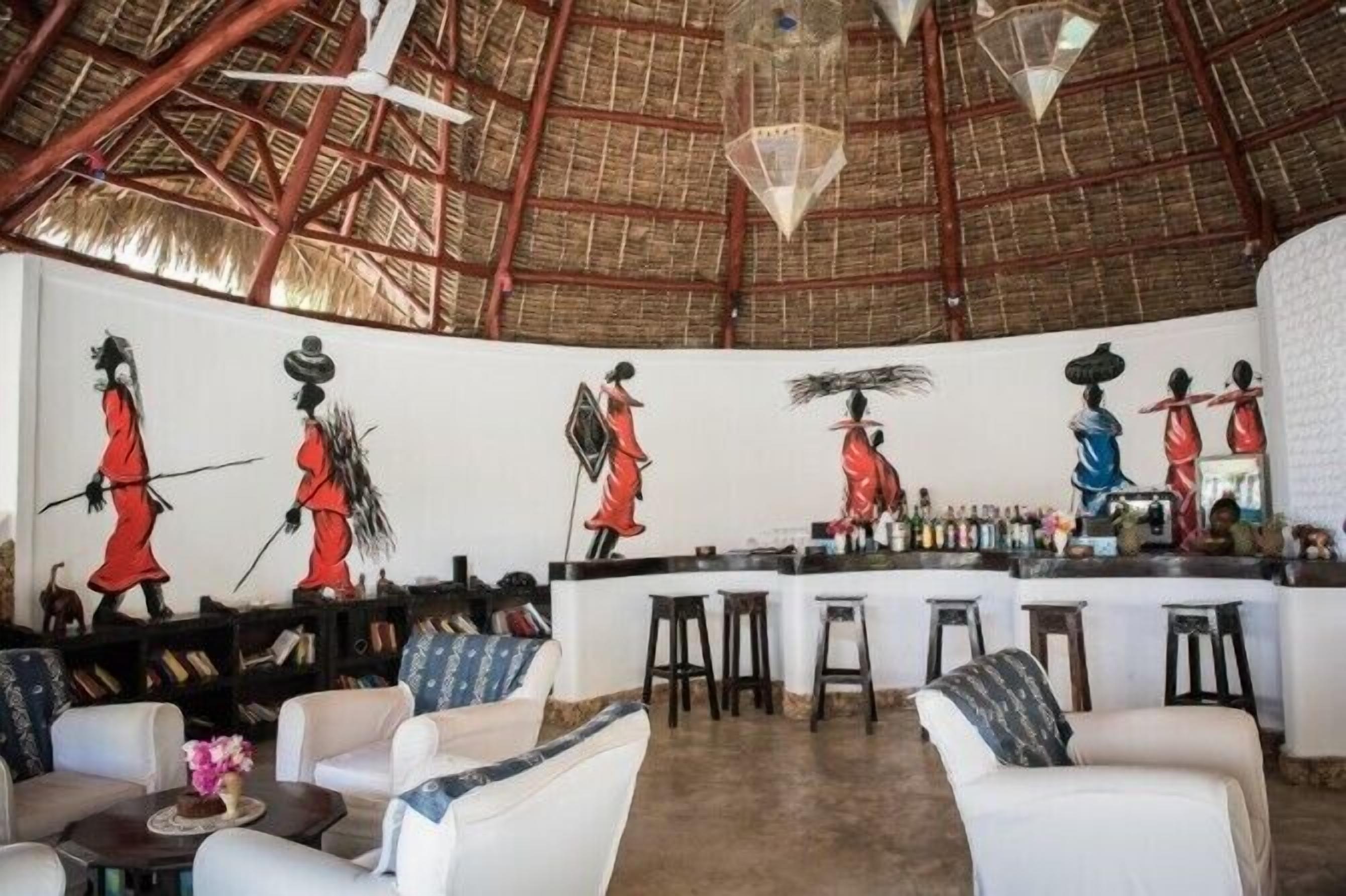 Angolo bar con soffitto alto e tetto in paglia, con un affresco raffigurante figure che trasportano oggetti. Sono presenti sgabelli da bar e una zona salotto con divani.