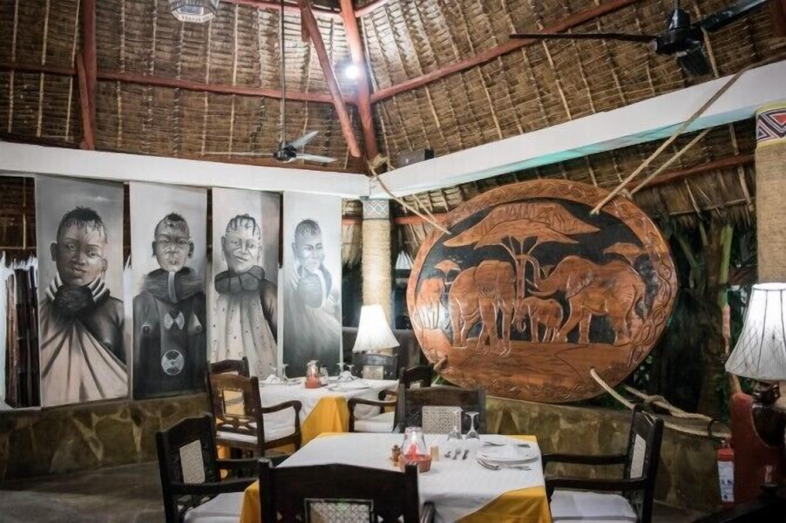 Ein Restaurant mit einem Strohdach, Holztischen und -stühlen sowie einem großen Wandgemälde von Elefanten.
