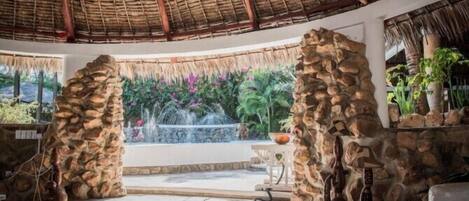 A circular room with a thatched roof, stone pillars, and a central fountain.