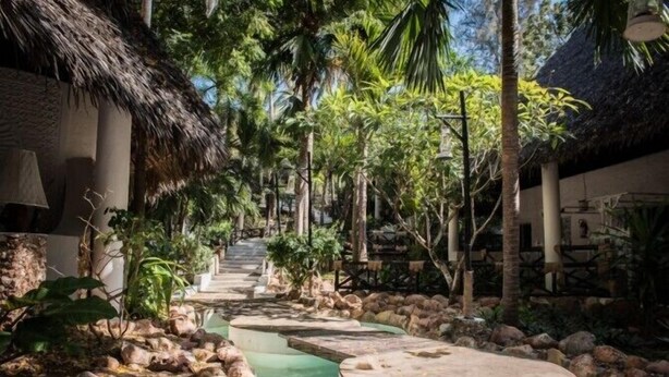A tropical resort pathway with thatched-roof buildings, surrounded by lush greenery and a water feature. - Scorpio Villas Resort (Malindi)