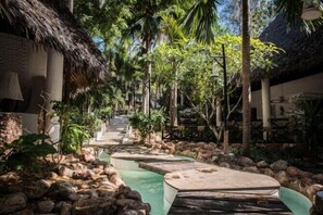 A tropical resort pathway with thatched-roof buildings, surrounded by lush greenery and a water feature. - Scorpio Villas Resort (Malindi)