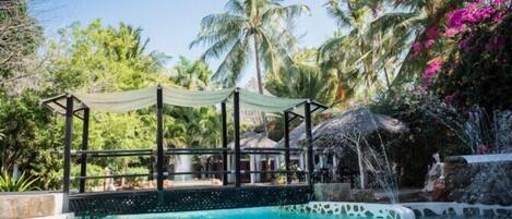 A pool area with a thatched-roof structure, surrounded by palm trees and a waterfall feature.