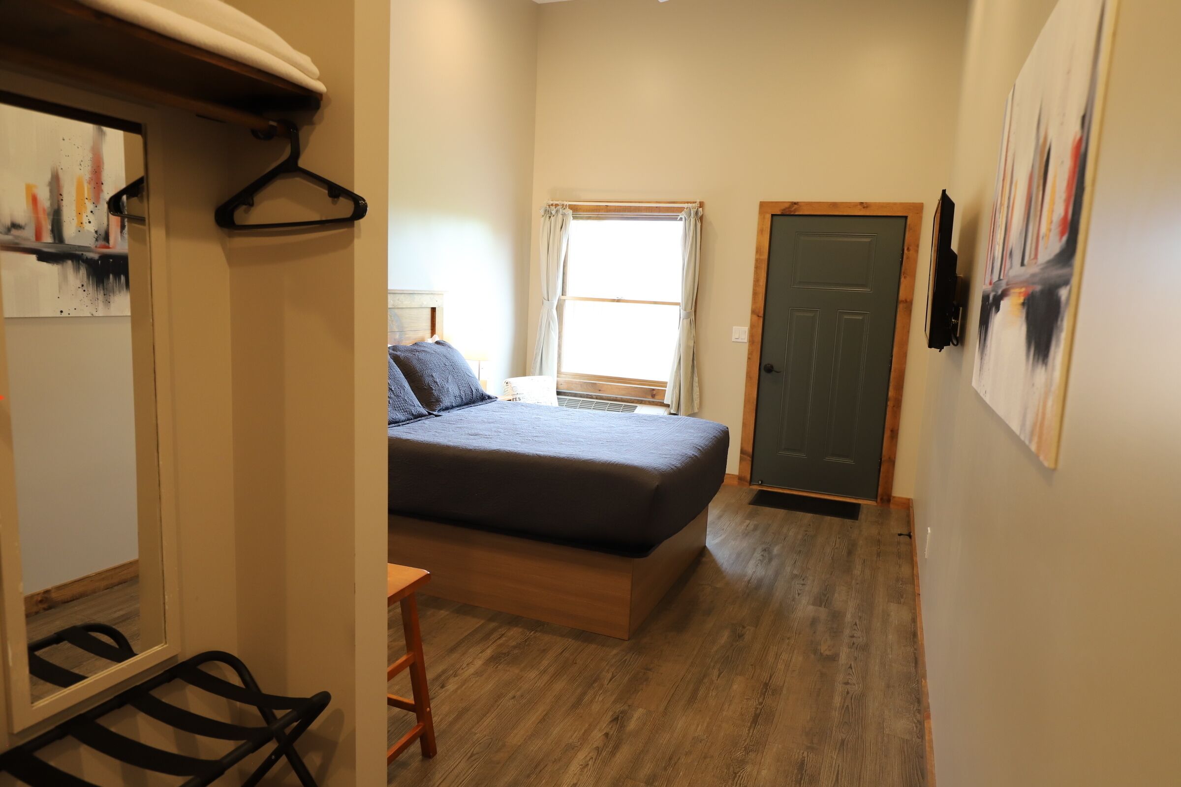 Un dormitorio con cama, suelo de madera, ventana con cortinas, puerta y televisor montado en la pared.