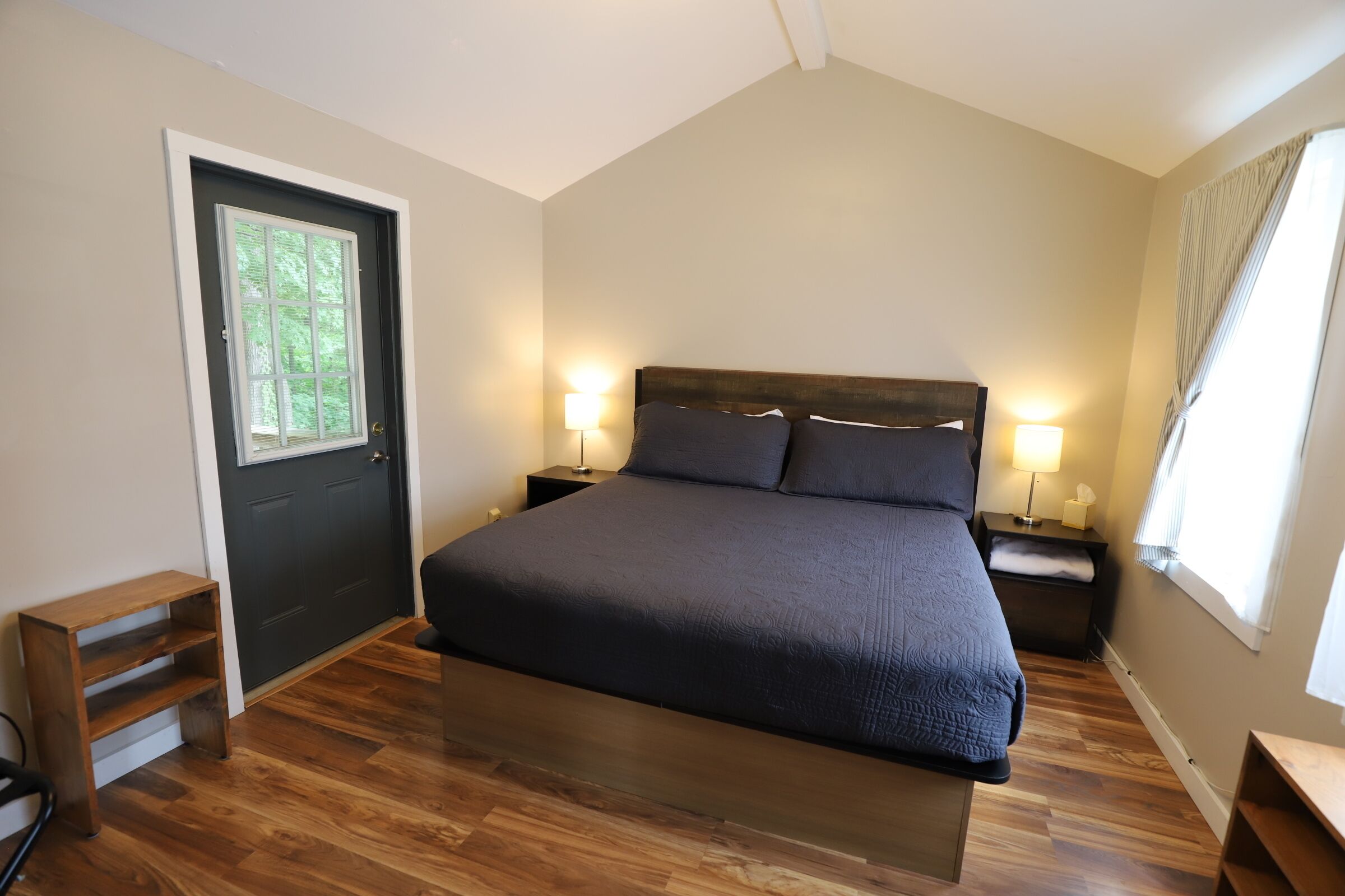 A bedroom with a large bed, two bedside tables with lamps, a wooden bench, a door with a window, and a wooden floor.