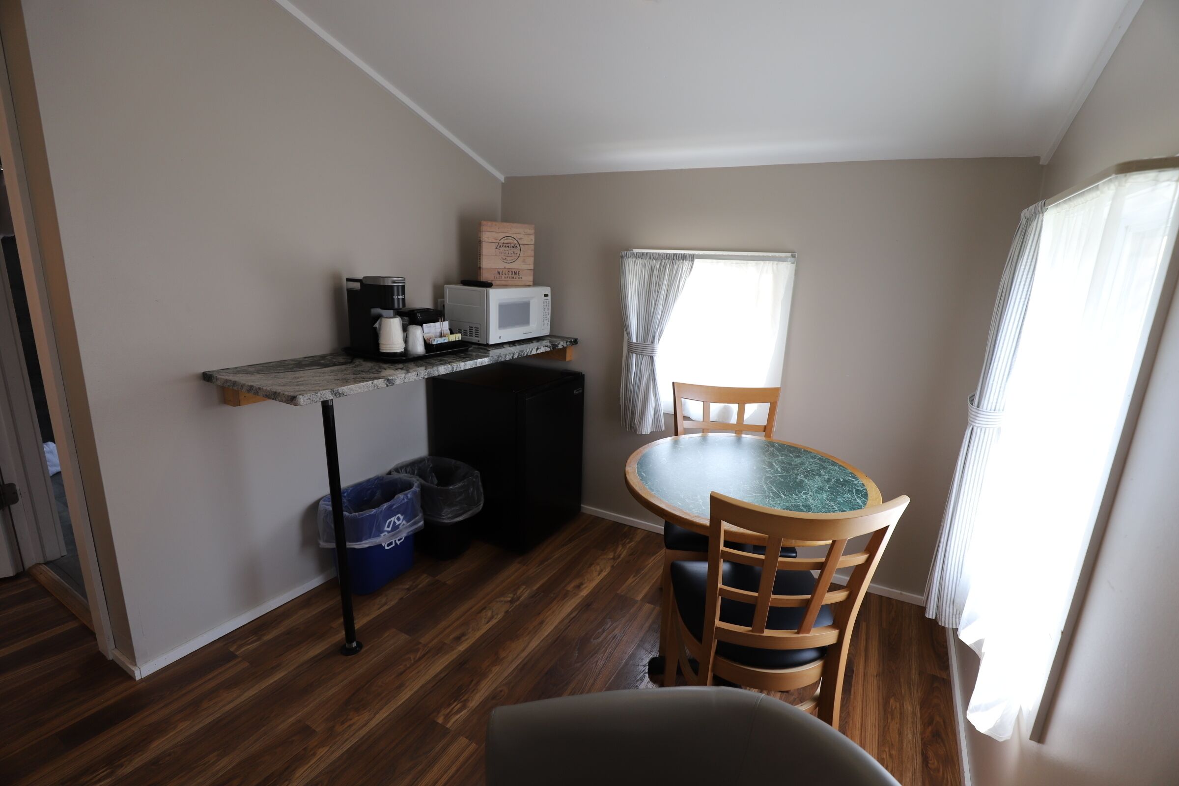 A compact kitchen with a microwave, coffee maker, and storage boxes. There is a dining table with chairs and a window with curtains.