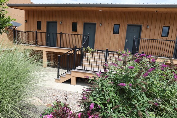 A wooden building with a balcony, two doors, and a small garden area with purple flowers.