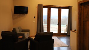 Una sala de estar con una televisión de pantalla plana, dos sillones, un sofá y un ventanal corredera que da a un espacio exterior.