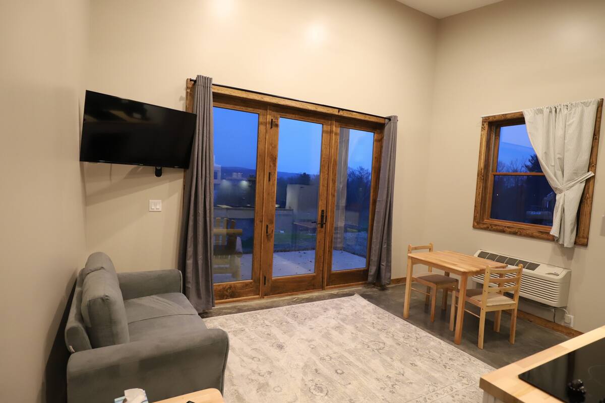 A living room with a sofa, a TV mounted on the wall, a dining table with chairs, and a large sliding glass door leading to an outdoor area.