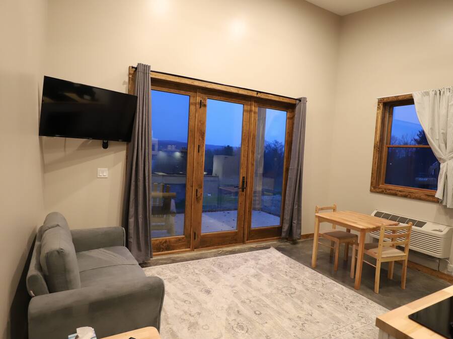 Una sala de estar con sofá, televisor montado en la pared, mesa de comedor con sillas y un amplio ventanal corredizo que da a un espacio exterior.