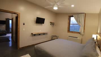 Un dormitorio con cama, ventilador de techo, televisión, ventana con cortinas y una pequeña estantería.