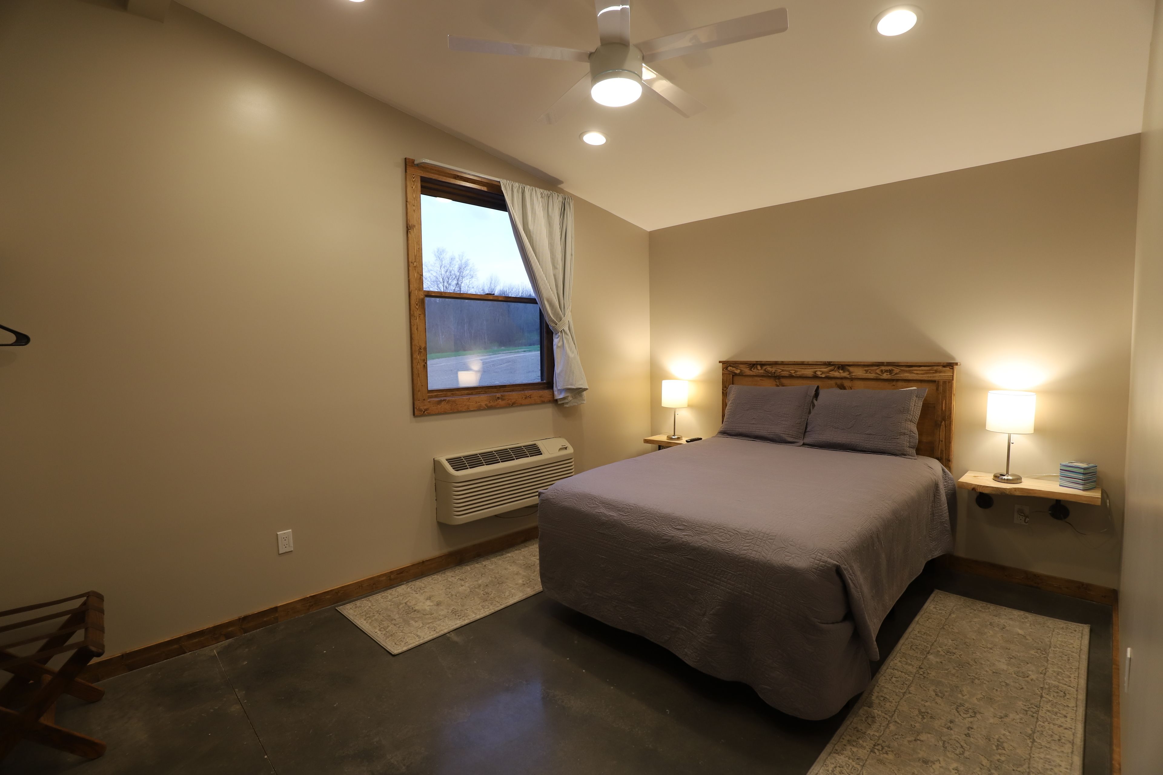 A bedroom with a bed, two bedside lamps, a ceiling fan, a window with curtains, a heater, and a small rug.