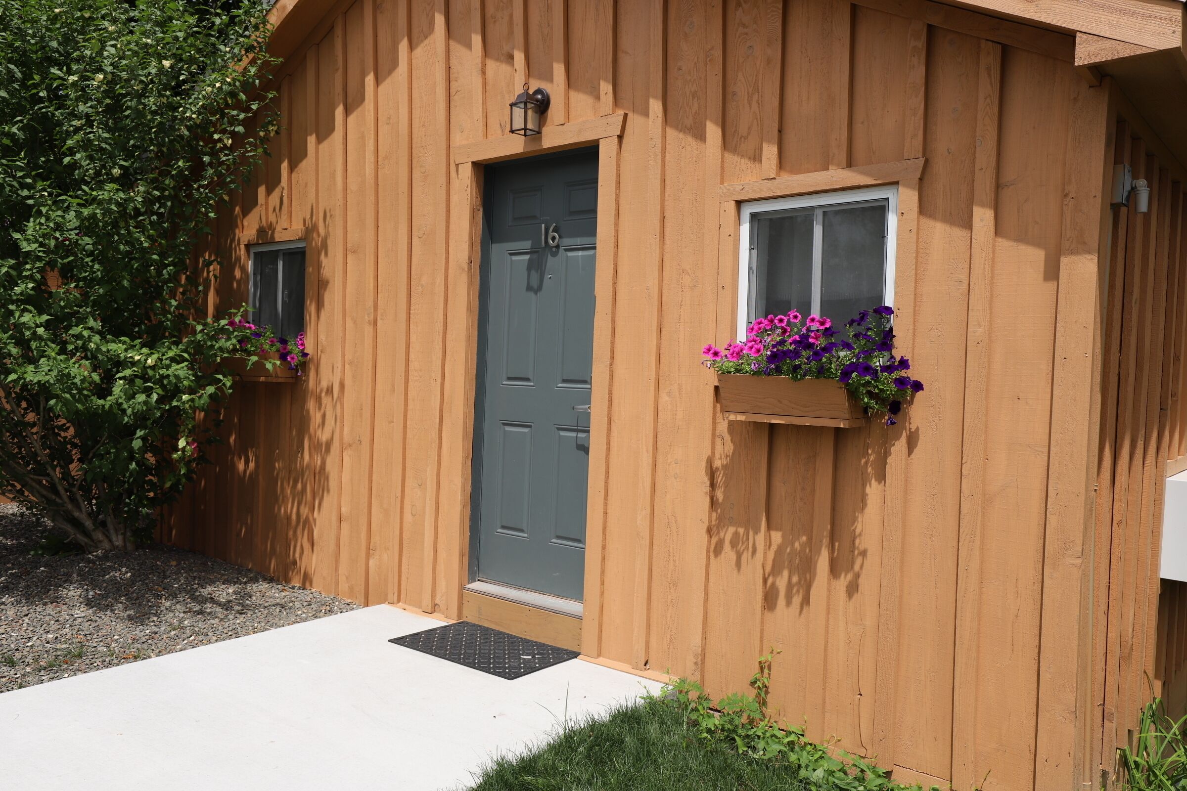 Un bâtiment en bois avec une porte grise, deux fenêtres ornées de jardinières et un petit porche.