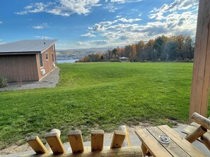 A wooden deck with a table and chairs, a grassy field, a building with a sloped roof, and a distant view of trees and a body of water. - Lakeside Resort (Watkins Glen)