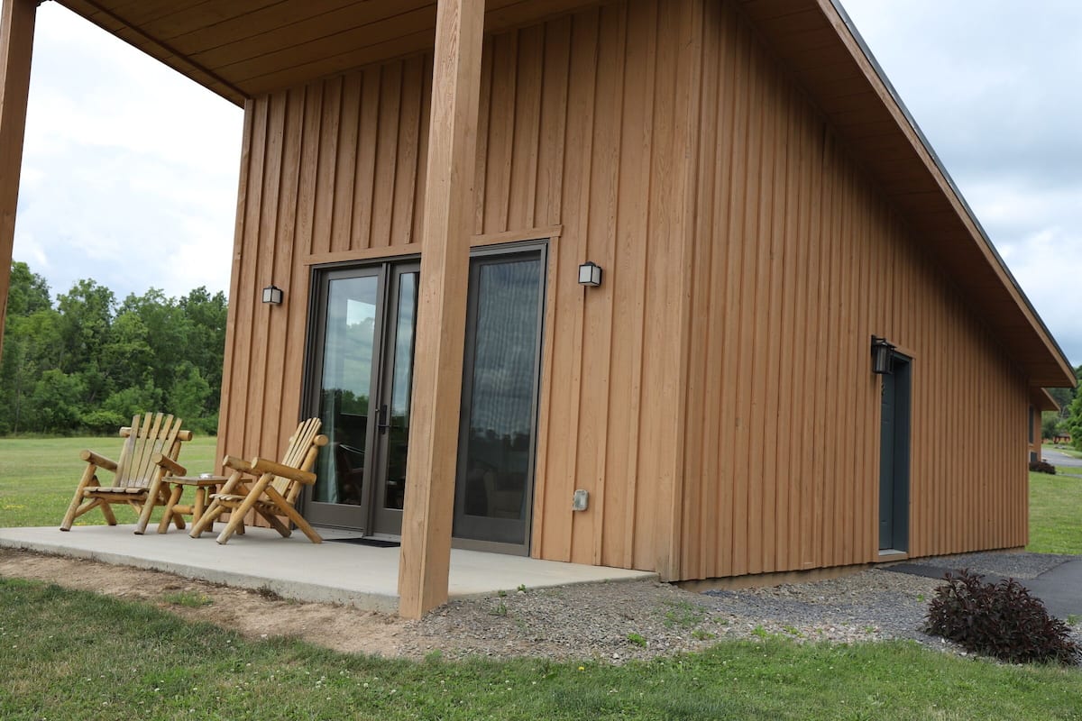 A wooden cabin with a covered porch, two Adirondack chairs, and a sliding glass door.