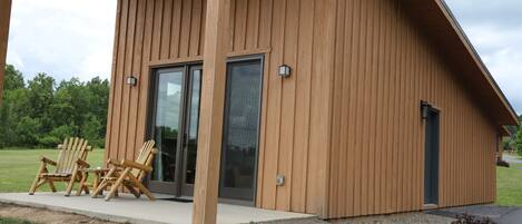 A wooden cabin with a covered porch, two Adirondack chairs, and a sliding glass door.