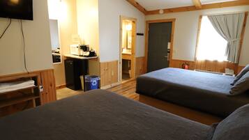 A hotel room with two beds, a TV mounted on the wall, a desk with a computer, and a window with curtains.