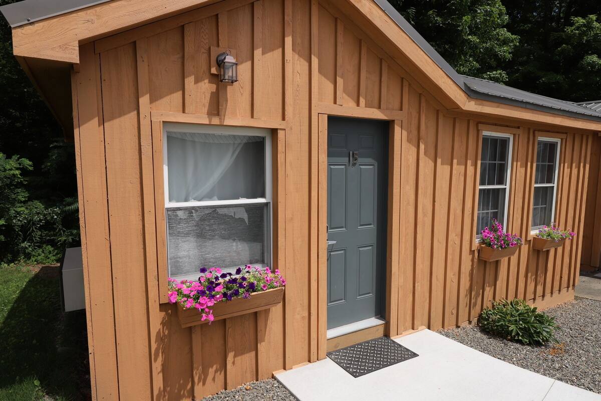 A wooden cabin with a gray door, a window with a curtain, and flower boxes on the windowsill.
