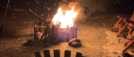 A chair facing a campfire with logs and a metal grill in the foreground, set in a snowy outdoor area.