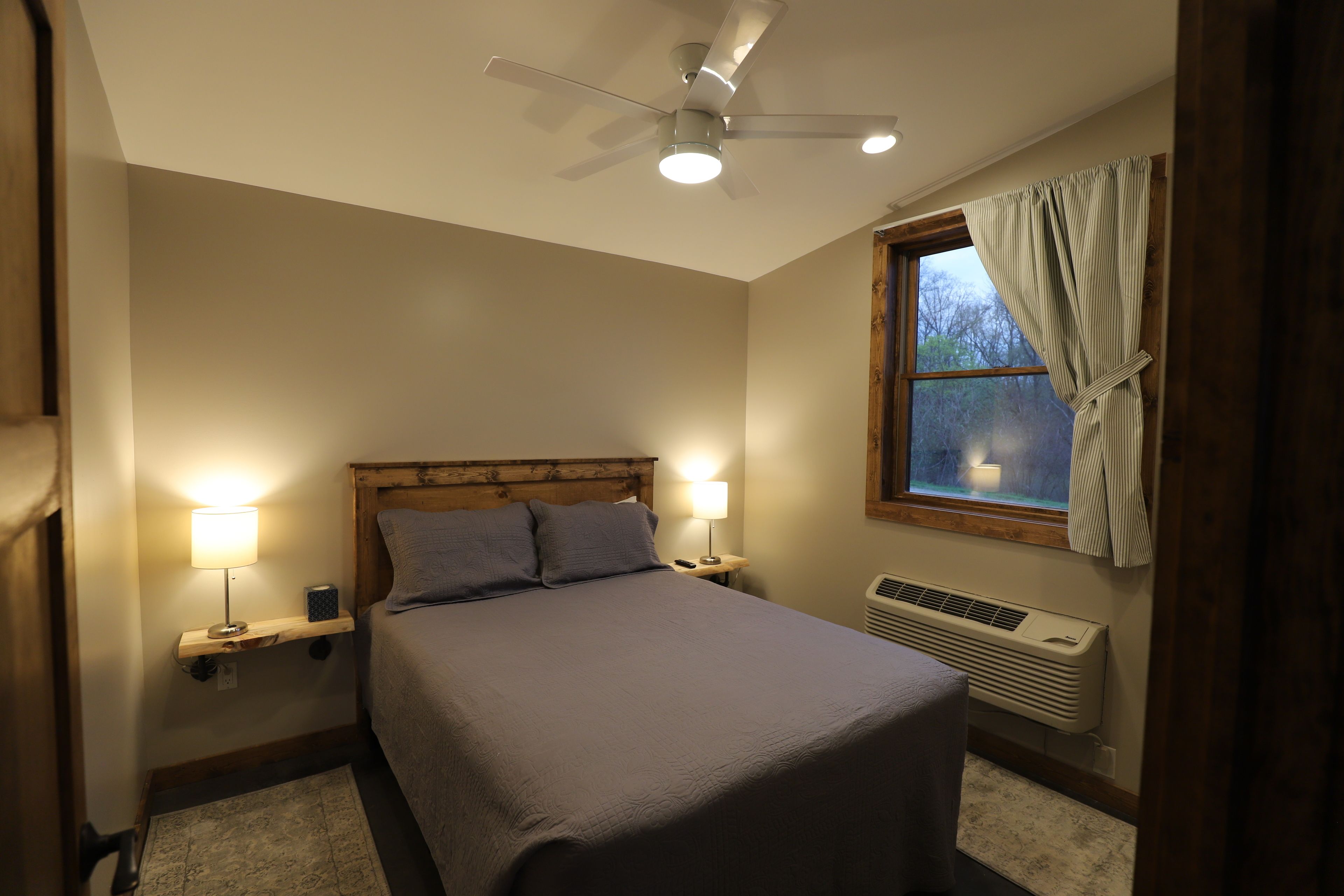 A bedroom with a bed, two bedside lamps, a ceiling fan, a window with curtains, and a wall-mounted heater.