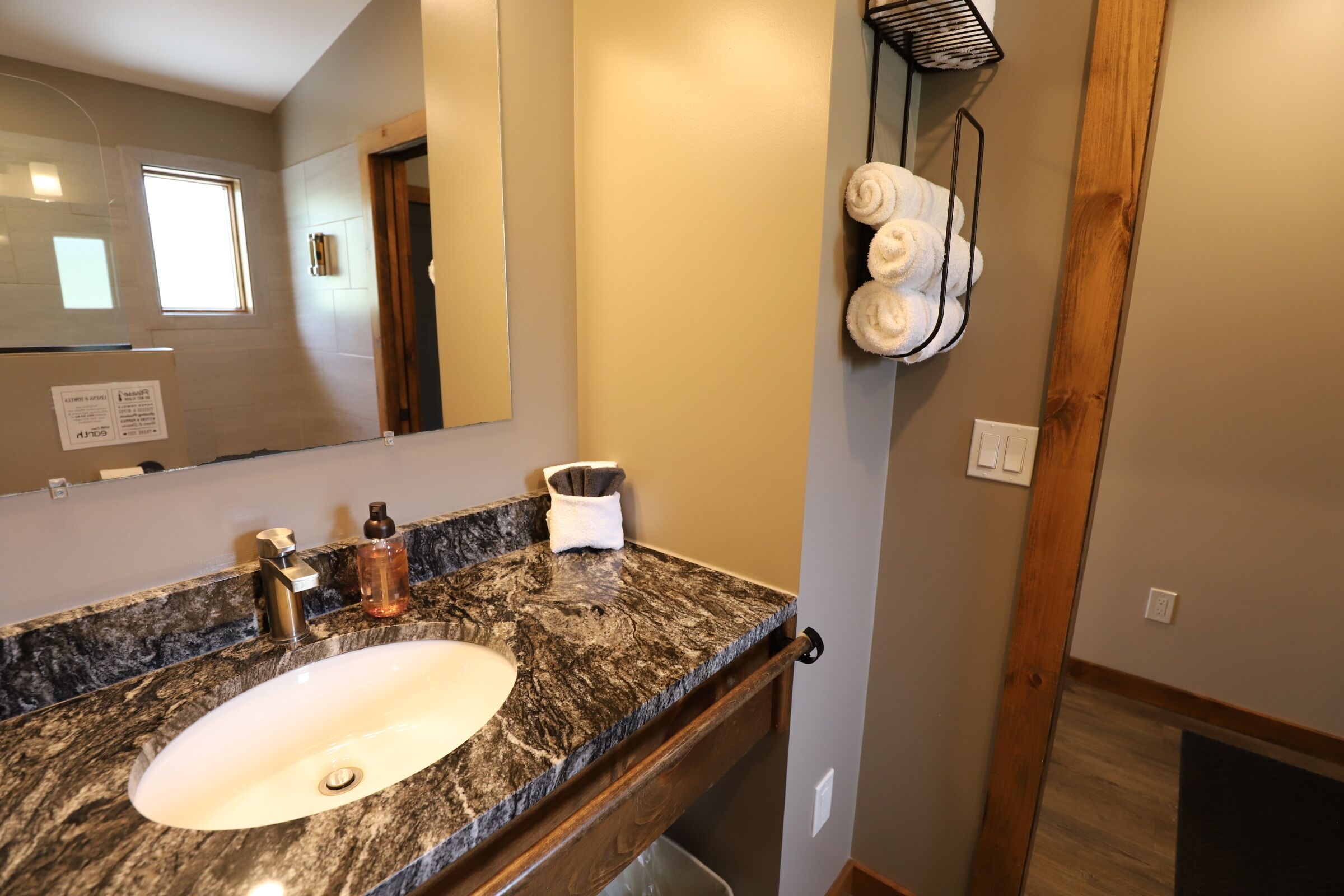 Une salle de bain avec un lavabo doté d’un plan de travail en granit, un miroir, un porte-serviettes avec des serviettes et une porte en bois.