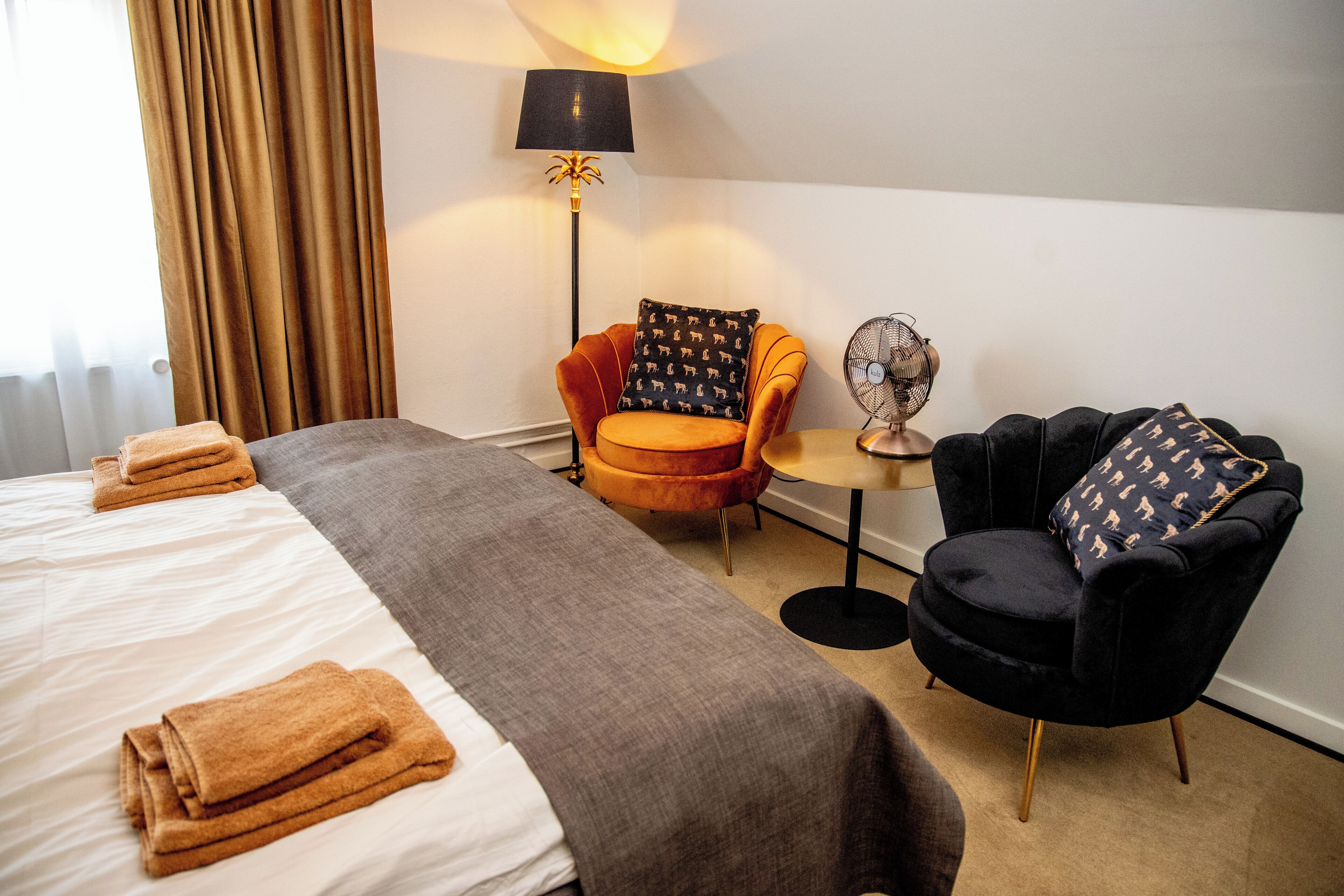 Una habitación de hotel con una cama, un sofá gris, un sillón naranja, un sillón negro, una lámpara de pie y una mesita.
