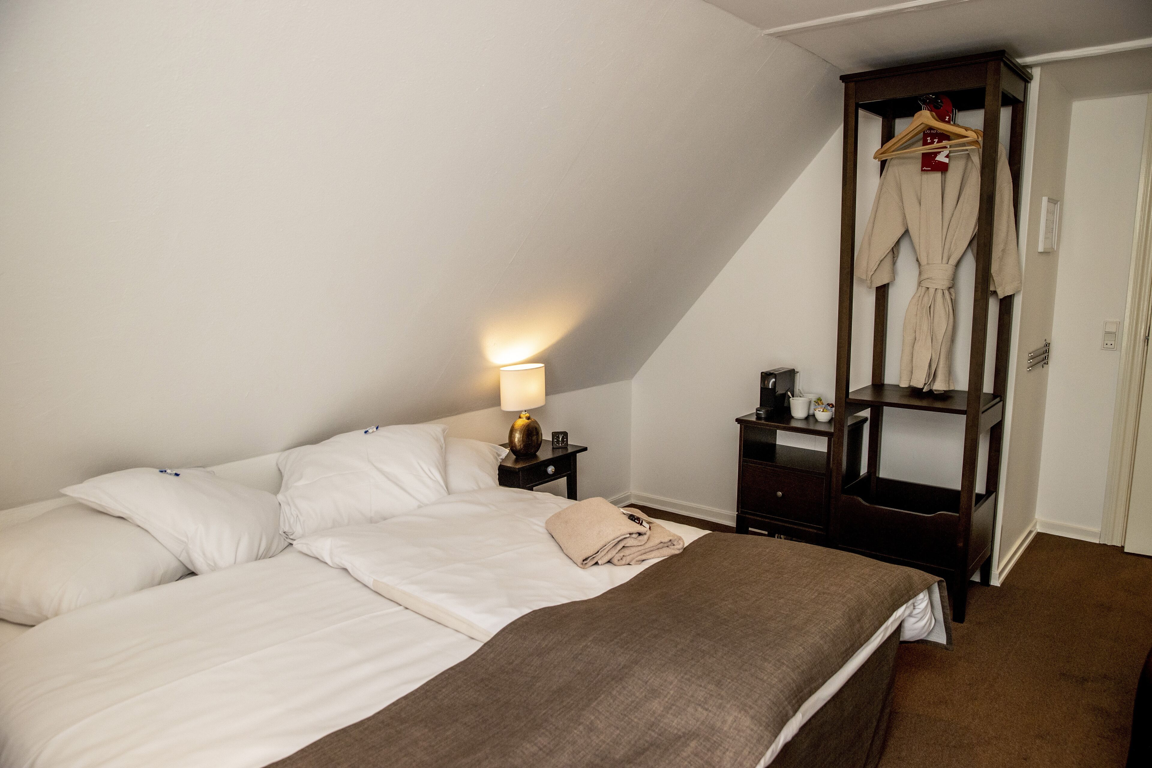 A neatly made bed with white linens, a bedside lamp, a wooden nightstand, and a clothing rack with a robe.