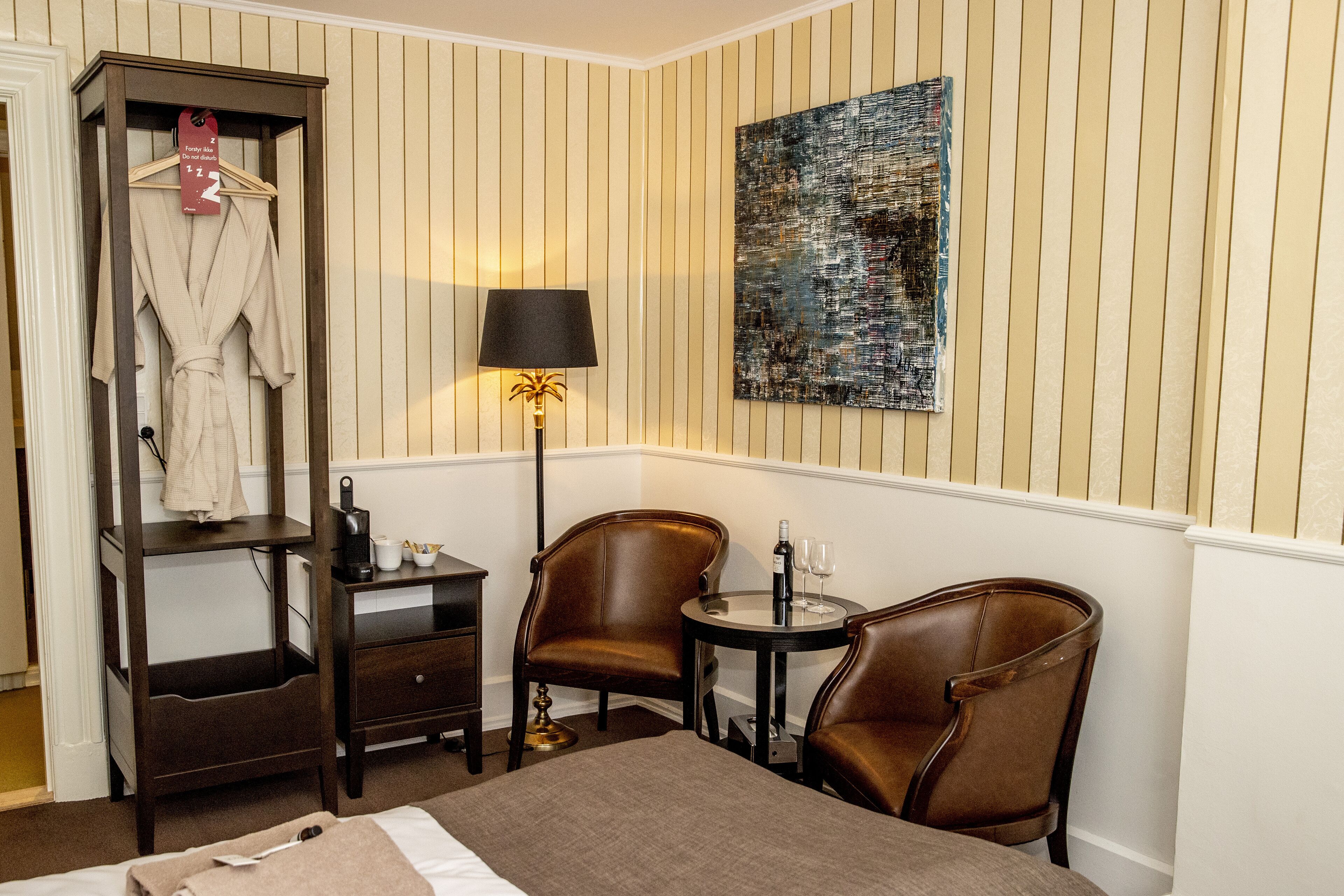 A hotel room with a bed, a robe hanging on a rack, a desk with a lamp, two chairs, and a small table with a bottle and glasses.