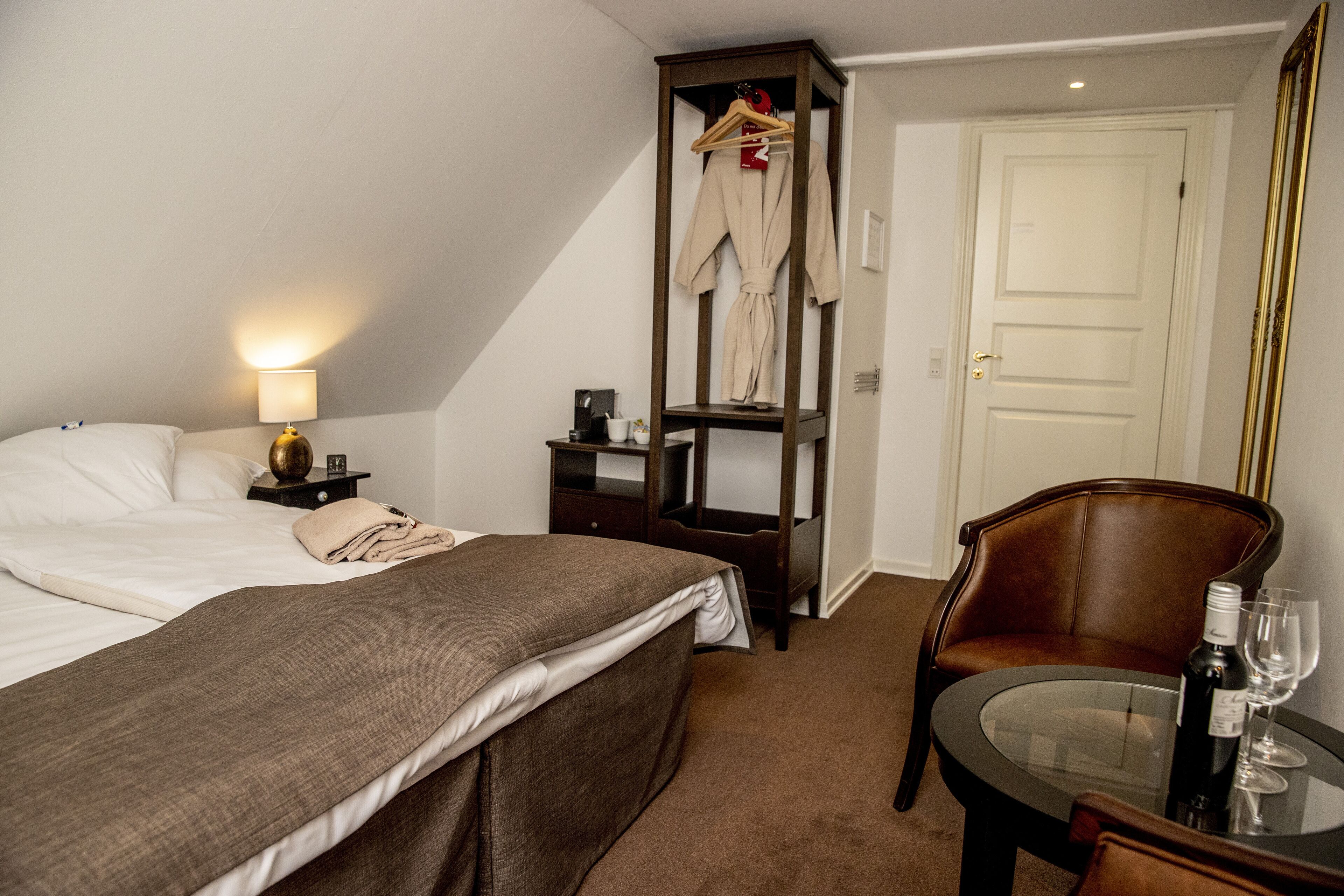 Une chambre d’hôtel comprenant un lit, une table de chevet avec une lampe, une chaise, une petite table avec une bouteille et des verres, et un porte-manteau avec un peignoir.